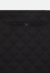 Czarna teksturowana powierzchnia z powtarzającymi się trójkątnymi logo orła Emporio Armani oraz centralnym metalowym napisem marki Emporio Armani i emblematem orła.