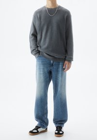 Grau gestrickter Pullover, lockere blaue Jeans mit ausgebleichten Bereichen und schwarz-weiße Sneakers. Silberne Kettenhalskette als bemerkenswertes Detail.