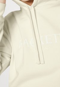 Hoodie w kolorze kremowym, z kieszenią z przodu, sznurkami oraz wytłoczonym białym napisem logo. Miękka tkanina i przytulny, luźny krój.
