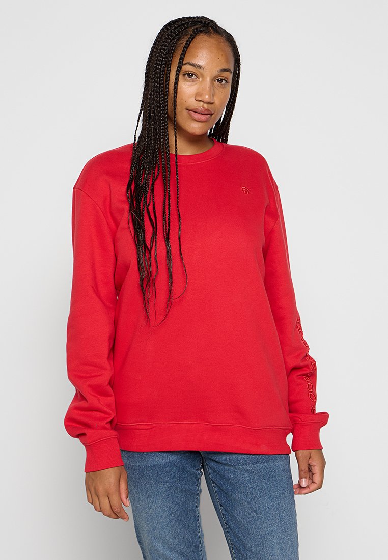 Röd sweatshirt med långa ärmar, rundad halsringning, ribbade muddar och nederkant. Har en liten logotyp på bröstet och text längs ärmen.
