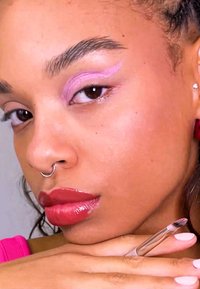 Close-up van een gezicht met roze oogschaduw, glanzende rode lippen en een zilveren septumring; het haar is golvend en de huid heeft een gladde textuur.