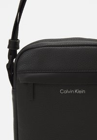 Borsa a tracolla in pelle nera con struttura a granulato, dotata di chiusura con zip, tracolla regolabile e logo "Calvin Klein" in argento.