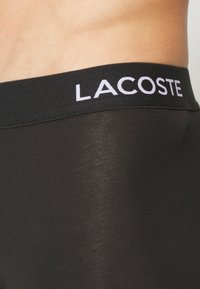 Black men's underwear featuring a smooth, stretchy fabric, with a wide elastic waistband displaying the "LACOSTE" logo in white.