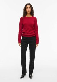Pull en tricot rouge avec des manches longues, associé à un pantalon noir à jambe droite et des bottes à talons noires, se tenant devant un fond blanc.