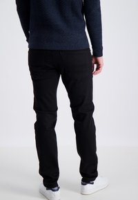 Jean noir slim, avec des poches arrière et des accents de couture subtils, associés à un pull marine texturé. Baskets blanches visibles.