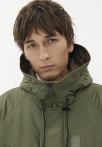 Olive zelena izolirana jakna z visokim ovratnikom, nastavljivimi vrvicami, zadrgo in subtilnimi šivi. Mehka, gladka tekstura.