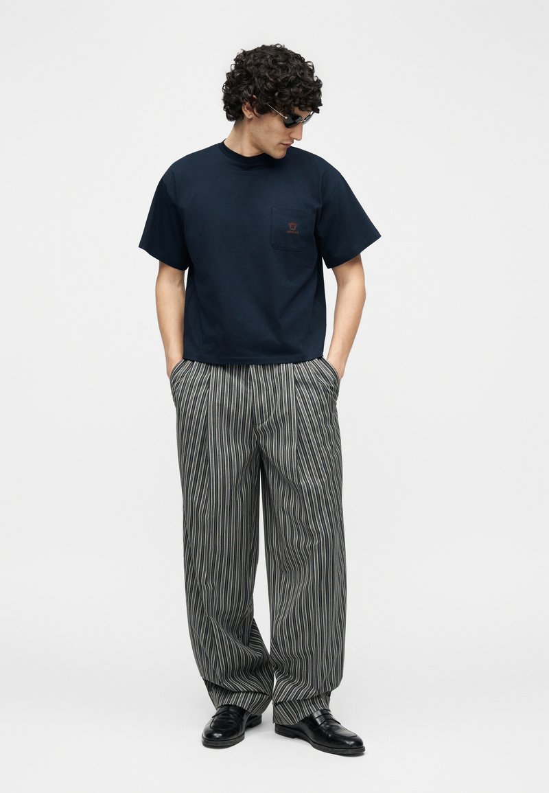 Uomo che indossa occhiali da sole neri, camicia blu scuro a maniche corte con taschino sul petto, pantaloni a righe larghe grigio e bianco, e scarpe nere, con le mani in tasca.