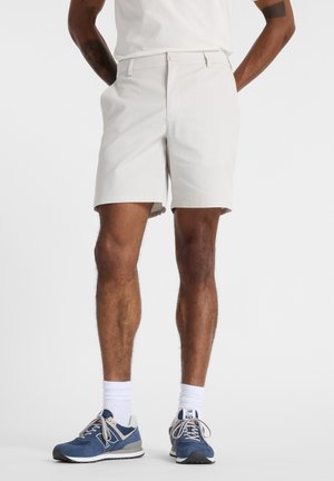 Mann trägt weiße Shorts bis zum Knie, weiße Sportsocken und blaue Sportschuhe, steht mit den Händen hinter dem Rücken.