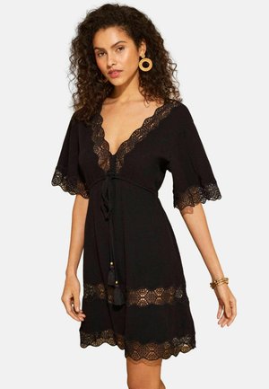 Schwarzes Kleid mit kurzen, ausgestellten Ärmeln, verziert mit Spitzenbesatz und einer Schnürung in der Taille. Akzente setzen Quasten und eine gezackte Spitze am Saum.