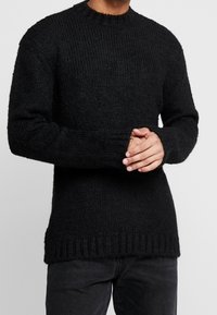 Svart stickad tröja med ribbad textur, rund halsringning och långa ärmar. Har en något längre bakdel och detaljerade manschetter.