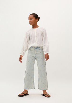 Camisa blanca texturizada de manga larga con botones, combinada con unos jeans de pierna ancha de color azul claro con bordado floral y un acabado desgastado. Sandalias negras.