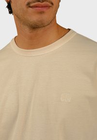 Beige puuvillane T-särk klassikalise ümarakaelusega, millel on väike tikandiga elevandi logo vasakul rinnal ja sile tekstuur.