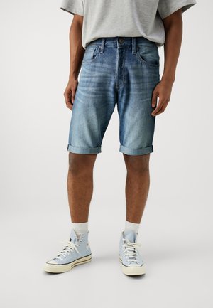 Mand iført lyseblå op rullede denimshorts, lysegrå t-shirt, hvide sokker og lyseblå høje sneakers, står på ensfarvet baggrund.