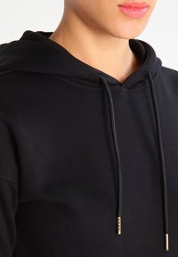 Felpa nera con cappuccio realizzata in tessuto morbido, dotata di tasca a marsupio, cordino regolabile con dettagli dorati e polsini a costine.