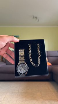 Zilveren roestvrijstalen horloge met zwarte wijzerplaat en chronograaffunctie naast een bijpassende zilveren ketting in een zwarte presentatiedoos.