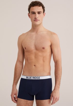 Mężczyzna bez koszulki, ubran w granatowe bokserki z białym pasem z napisem „BLUE RIDGE”, stojący na jasnym, jednolitym tle.