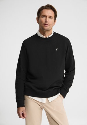 Hombre que lleva un suéter negro sin cuello sobre una camisa blanca de botones y pantalones beige, de pie con una mano en el bolsillo.