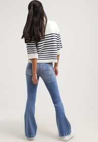 Kvinna med långt mörkt hår klädd i en vit och marinblå randig tröja, ljusblå utsvängda jeans och vita sneakers, stående vänd bort.