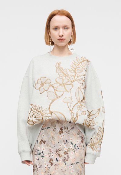Γυναίκα με κοντά κόκκινα μαλλιά φοράει λευκό υπερμεγέθες φούτερ με χρυσή κεντημένη λουλουδένια διακόσμηση και μπεζ φλοράλ φούστα, κοιτώντας προς τα εμπρός.