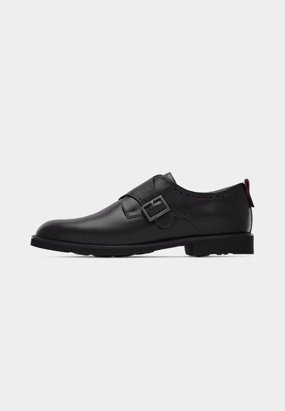 Chaussure habillée en cuir noir pour homme, avec une seule bride à boucle argentée et un petit talon, sur un fond clair uni.