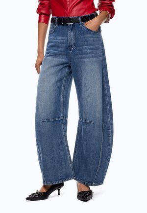 Donna che indossa jeans blu a gamba larga, top rosso a maniche lunghe, cintura nera e scarpe con tacco nere con dettagli decorativi.