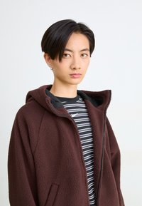 Chaqueta de forro polar marrón con capucha y cierre de cremallera, llevada sobre una camisa de rayas en blanco y negro. La chaqueta tiene bolsillos laterales y una textura suave.