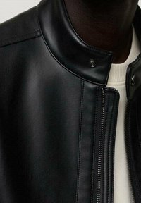 Czarna skórzana kurtka z wysokim kołnierzem, zapięciem na zatrzaski i bocznym suwakiem. Gładka faktura z minimalną ilością przeszyć.