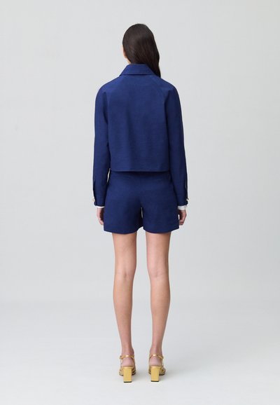 Tenue en lin bleu marine composée d'une veste courte et d'un short avec une coupe décontractée, mettant en avant des poignets boutonnés et des poches, portée avec des sandales à talons jaunes.