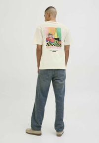 Beige bomulds t-shirt med et farverigt grafik af et stilleben med en flaske og frugt. Løs pasform, korte ærmer, set bagfra, parret med blå jeans.