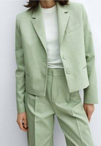 Vrouw in een lichtgroen maatpak met een cropped blazer en hooggesneden geplooide broek, gecombineerd met een witte getailleerde blouse.