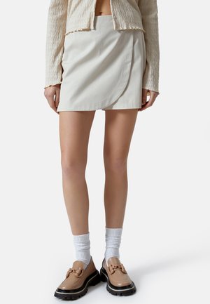 Gonna avvolgente beige in tessuto liscio, caratterizzata da un design con cuciture curve. Abbinata a scarpe beige chiare e calze bianche a coste.