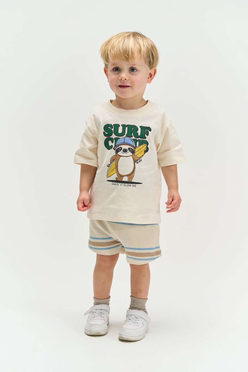 Tout-petit aux cheveux blonds portant un T-shirt crème avec un motif de paresseux surfeur et un short rayé, debout devant un fond uni.