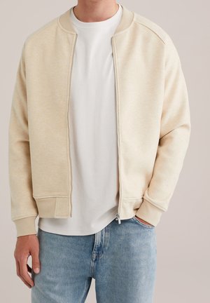 Mann trägt beiger Zipper-Jacke über weißem T-Shirt und hellblauen Jeans, mit einer Hand in der Tasche und sichtbarem Reißverschlussdetail.