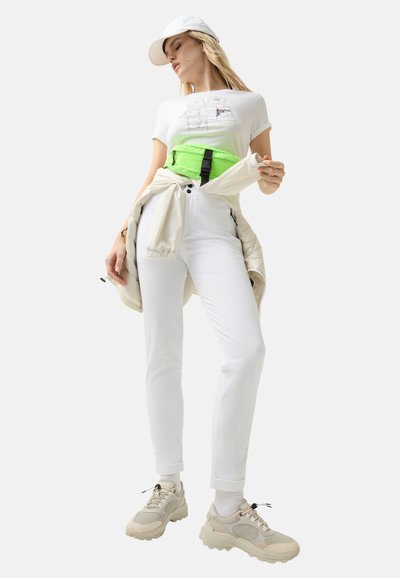 White fitted pants, white graphic t-shirt, neon green waist bag with black hardware, beige sneakers, and a white cap. Casual urban outfit.
