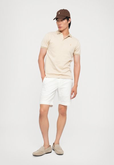Beige polo skjorte med korte ermer, hvite shorts og lyse grå loafers. Brun caps og uformell positur mot en enkel bakgrunn.