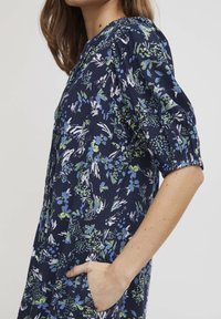 Mornarska cvetna obleka z zelenimi in modrimi vzorci, kratkimi naborki na rokavih in stranskimi žepi. Tkanina se zdi lahka in ima gladko teksturo.