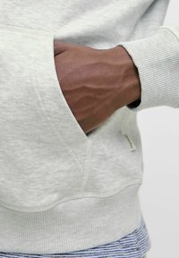 Hanorac gri deschis cu buzunar tip kengur, manșete rib variant și o textură moale. Detaliu al unei mâini în buzunar, evidențiind țesătura și construcția.