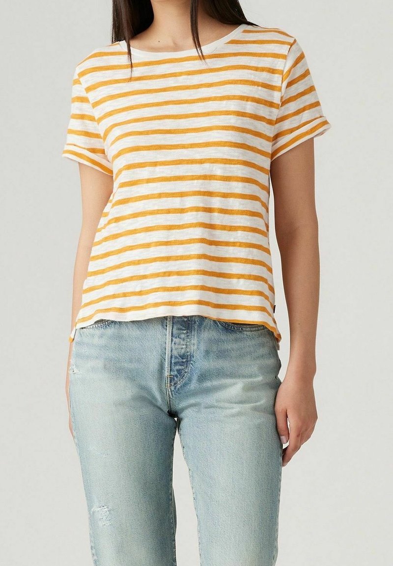 Femme portant un t-shirt à manches courtes rayé jaune et blanc avec un jean bleu clair, debout devant un fond uni clair.