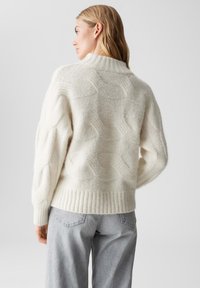 Maglione color crema con un design a trecce testurizzato. Vestibilità comoda con colletto alto e orlo a coste, indossato con pantaloni grigio chiaro.