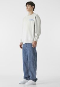 Világosszürke pulóver kék felirattal, laza szabású. Laza farmernadrággal és fehér tornacipővel párosítva, hangsúlyozva a laza utcai stílust.