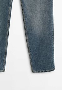 Section de jambe de jean en denim bleu avec coutures latérales visibles et légère décoloration, présentée sur un fond blanc.