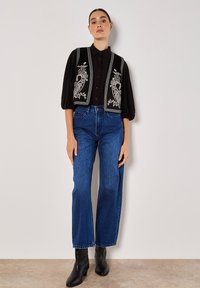 Schwarze Button-Up-Hemd kombiniert mit einer bestickten Weste mit Blumenmustern. Blaue hoch taillierte, weite Jeans und schwarze Stiefeletten.