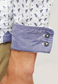 Gros plan d'une manche de chemise blanche retroussée avec des motifs floraux bleus et un poignet bleu uni fermé par deux boutons sur le bras d'une personne.
