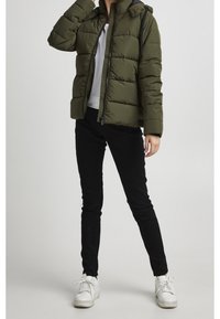 Olivgrön vadderad jacka med huva, försedd med dragkedja och ribbade muddar, kombinerad med svarta slim fit-byxor och vita sneakers.