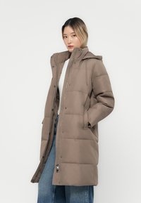 Manteau rembourré marron avec fermeture éclair sur le devant, capuche et poches latérales. Présente un motif matelassé et un cordon de serrage au niveau du cou. Tissu texturé.