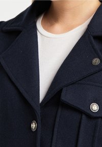 Detailní záběr na tmavě modrý kabát s kovovými knoflíky ve tvaru kotvy, nošený přes bílý crew-neck tričko, se zaměřením na límec a horní část hrudníku.