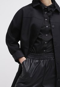 Černá oversized košile s kapsou na prsou, vyrobená z hladké tkaniny, s zapínáním na knoflíky, spárována s černými elastickými kalhotami z umělé kůže.