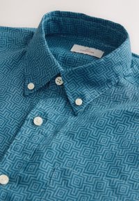 Camisa abotonada en verde azulado con un patrón geométrico. Cuenta con un cuello abierto y botones blancos. Está hecha de tela texturizada que muestra detalles finos.