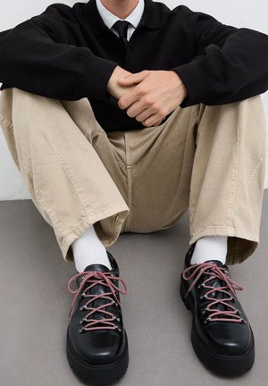 Black leather shoes with red and white patterned laces, thick soles, and metal eyelets. Worn with beige trousers and white socks.