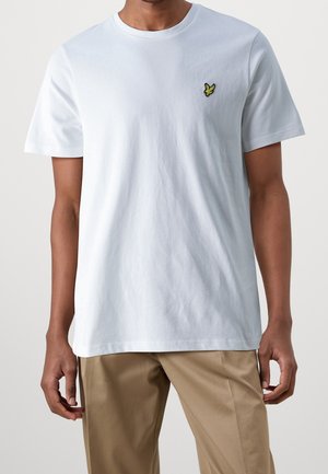Man draagt een eenvoudige witte T-shirt met korte mouwen en een klein geel vogellogo op de borst, gecombineerd met beige broek tegen een witte achtergrond.
